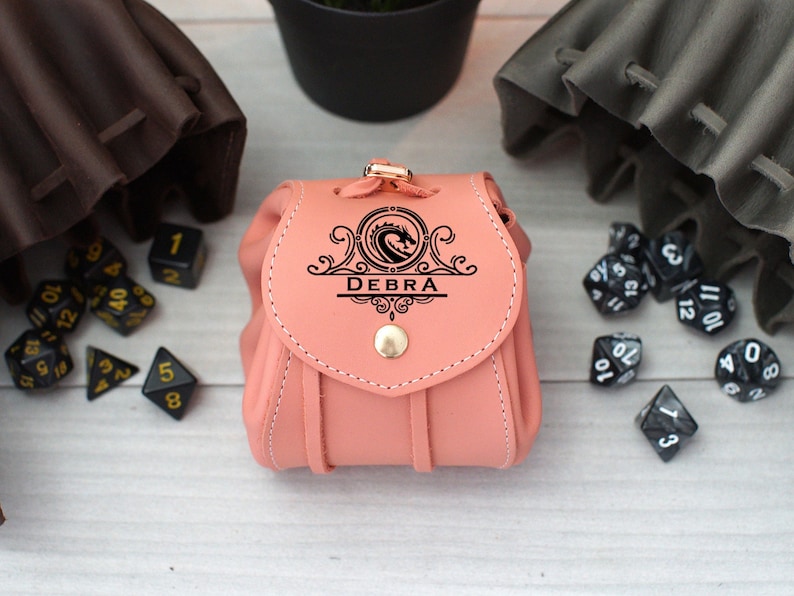 Pode incluir: Uma bolsa de dados de couro rosa com um design de drag&atilde;o e o nome "DEBRA" gravado na frente. A bolsa &eacute; cercada por dados pretos.