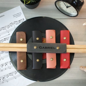 May include: A set of six leather drum stick holders with a personalized name tag that reads "GABRIEL". The holders are in brown, black, pink, and red.