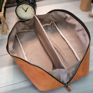 May include: Open tan leather travel case with a black zipper and interior dividers. The case is open, revealing a light tan interior with two compartments. The zipper pull is also tan leather.