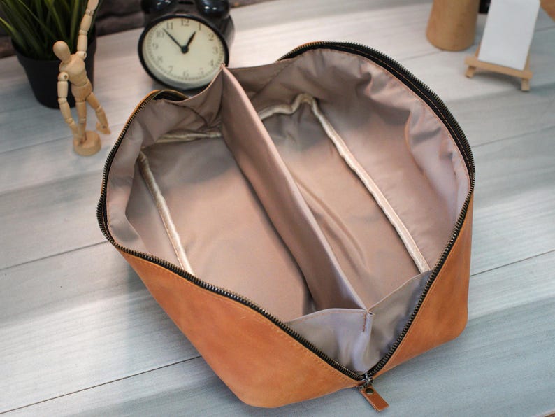May include: Open tan leather travel case with a black zipper and interior dividers. The case is open, revealing a light tan interior with two compartments. The zipper pull is also tan leather.