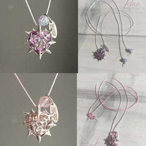 May include: Two necklaces with a silver heart pendant with spikes and a pink or purple crystal heart. The pendant has a small tag that says "Tears & Love". The necklaces are on a white or pink cord.