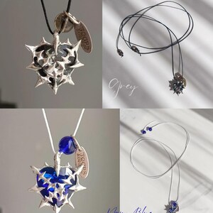 May include: A silver heart-shaped pendant necklace with a blue glass gem in the center. The pendant is surrounded by silver spikes and has a small silver tag that says "Tears & Beads". The necklace is available in grey and navy blue.