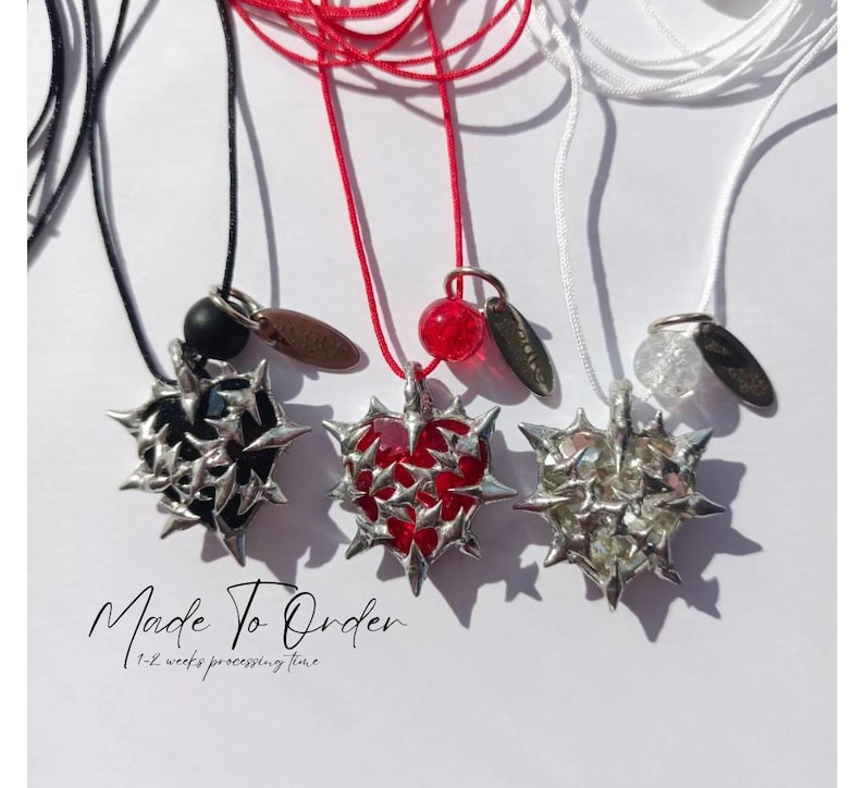 May include: Three necklaces with silver heart pendants featuring a thorny design. The hearts are filled with black, red, and clear resin. The necklaces are on black, red, and white cords. The text "Made To Order" and "1-2 weeks processing time" is below the necklaces.