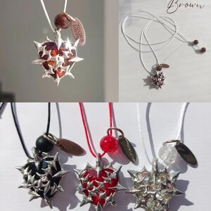 May include: A set of five necklaces with silver heart pendants featuring a thorny design. Each pendant has a different colored stone inside: brown, red, black, clear, and red. The necklaces are on white, black, and red cords. The tag on the necklace reads "Tears & Laughter".