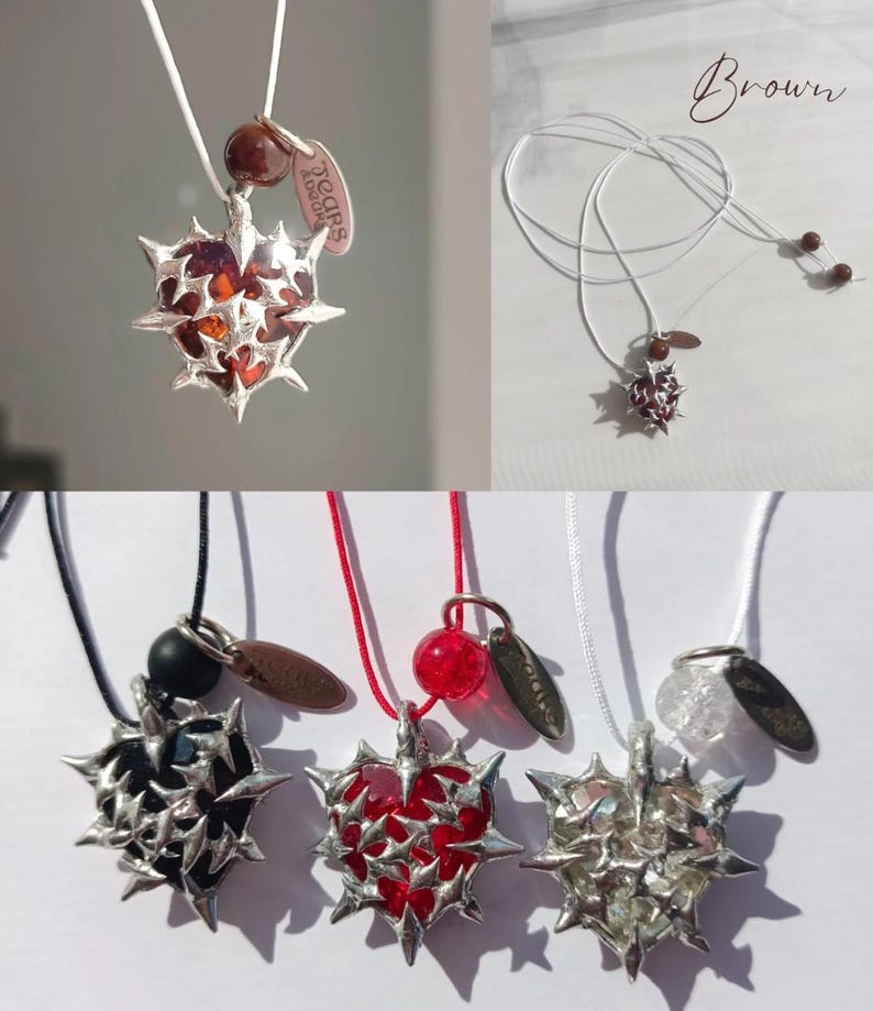 May include: A set of five necklaces with silver heart pendants featuring a thorny design. Each pendant has a different colored stone inside: brown, red, black, clear, and red. The necklaces are on white, black, and red cords. The tag on the necklace reads "Tears & Laughter".
