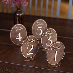 Puede incluir: Señales de números de mesa de madera con letras blancas, numeradas del 1 al 5. Los letreros ovalados están colocados sobre una mesa de madera oscura. Un jarrón con flores rosas está en el fondo.