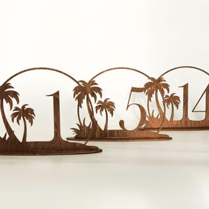 Puede incluir: Tres números de mesa de madera con temática tropical. Cada número, 1, 5 y 14, se establece contra un telón de fondo de palmeras y un marco circular. Los números y el paisaje están cortados de madera marrón oscuro.