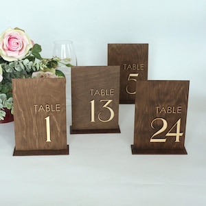 May include: Four wooden table number signs with gold lettering. The numbers are 1, 5, 13, and 24. The signs are dark brown with a rectangular shape and a base for standing. A bouquet of pink roses and greenery is in the background.