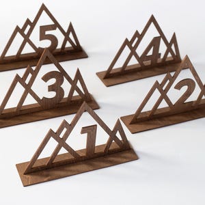 Könnte beinhalten: Fünf Tischkartenhalter aus Holz im Bergdesign. Jede Zahl, von eins bis fünf, ist aus dem Holz ausgeschnitten. Die Zahlen sind dunkelbraun und stehen auf weißem Hintergrund.