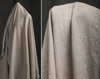 May include: A beige textured fabric with a crinkled appearance. The fabric is draped over a form, showcasing its soft and flowing texture.