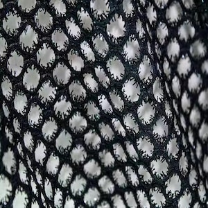 May include: Close-up of black mesh fabric with large, evenly spaced holes. The fabric has a textured appearance, with a slightly shiny finish. The background is a bright, off-white color, creating a contrast with the dark mesh.