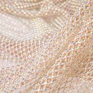 May include: Close-up of a shimmering, beige fabric with a net-like pattern. The fabric is densely covered with sparkling, iridescent rhinestones, creating a dazzling effect. The texture is soft and flowing, suggesting a luxurious material.