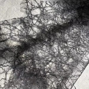 Puede incluir: Primer plano de una tela gris oscuro y transparente con un patrón complejo en forma de telaraña. La tela parece delicada y texturizada, con un aspecto ligeramente arrugado. El material probablemente se utiliza con fines decorativos.