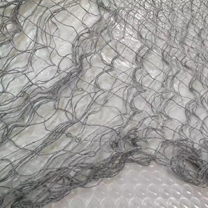 May include: Close-up of a gray fishing net with a textured appearance. The net is made of interwoven strands, creating a grid-like pattern. The net's color is a muted gray, and it appears to be made of natural fibers.