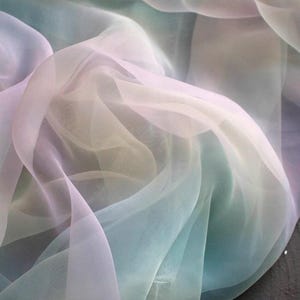 May include: A close-up of flowing, sheer fabric in a gradient of pastel colors. The fabric transitions from pale lavender to soft yellow and then to a light teal, creating a dreamy, ethereal effect. The material appears lightweight and delicate.