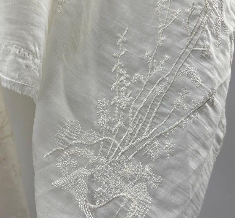 K&ouml;nnte beinhalten: Wei&szlig;er Baumwollstoff mit zarten wei&szlig;en Stickereien von Blumendesigns. Die Stickerei ist filigran und detailliert und erzeugt ein wundersch&ouml;nes und elegantes Muster.