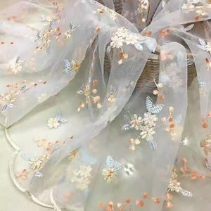 May include: Sheer white fabric with embroidered floral and butterfly designs in pastel colors. The delicate embroidery features light blue, peach, and cream-colored flowers and butterflies. The fabric is draped, creating soft folds and a flowing appearance.