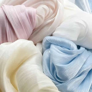 Könnte beinhalten: Eine Ansammlung weicher, durchscheinender Stoffstücke in Pastelltönen wie Hellrosa, Hellblau, Creme und Weiß. Das Material weist eine feine, gerippte Textur mit leichtem Glanz auf und ist in lockeren, überlappenden Falten und Schichten angeordnet.