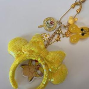 May include: A gold-toned safety pin keychain featuring a yellow sequined mouse ear headband charm, a yellow translucent mouse-shaped charm, a small star charm, and a circular charm with a rose graphic, all attached by gold-colored metal chains.