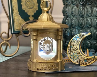 May include: A gold-colored lantern with a crescent moon design and the text "Ramadan Mubarak" on the base. The lantern is lit and has a white glow. The lantern is sitting on a table with a green book and a vase of flowers in the background.