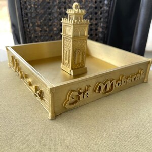 May include: A gold-colored tray with a small gold-colored mosque-shaped ornament in the center. The tray has the words "Eid Mubarak" written on the front.