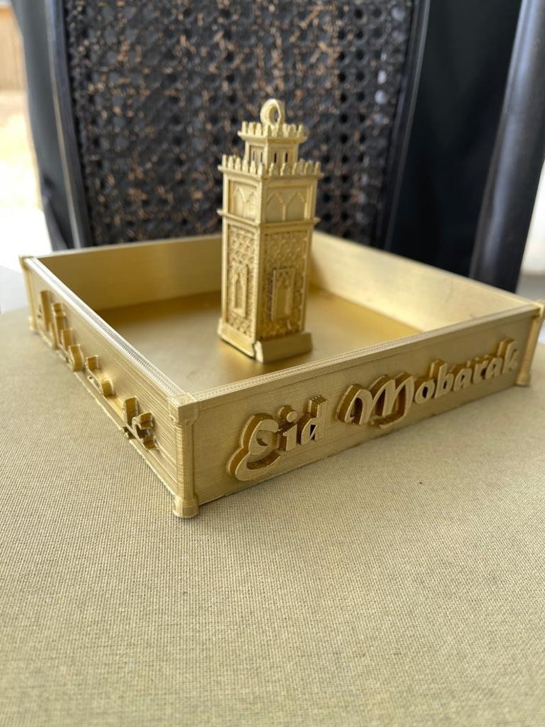 May include: A gold-colored tray with a small gold-colored mosque-shaped ornament in the center. The tray has the words "Eid Mubarak" written on the front.
