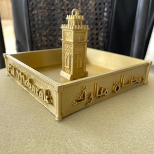 May include: A gold-colored tray with the words "Eid Mubarak" and Arabic script on the sides. A small gold-colored model of a minaret sits in the center of the tray.