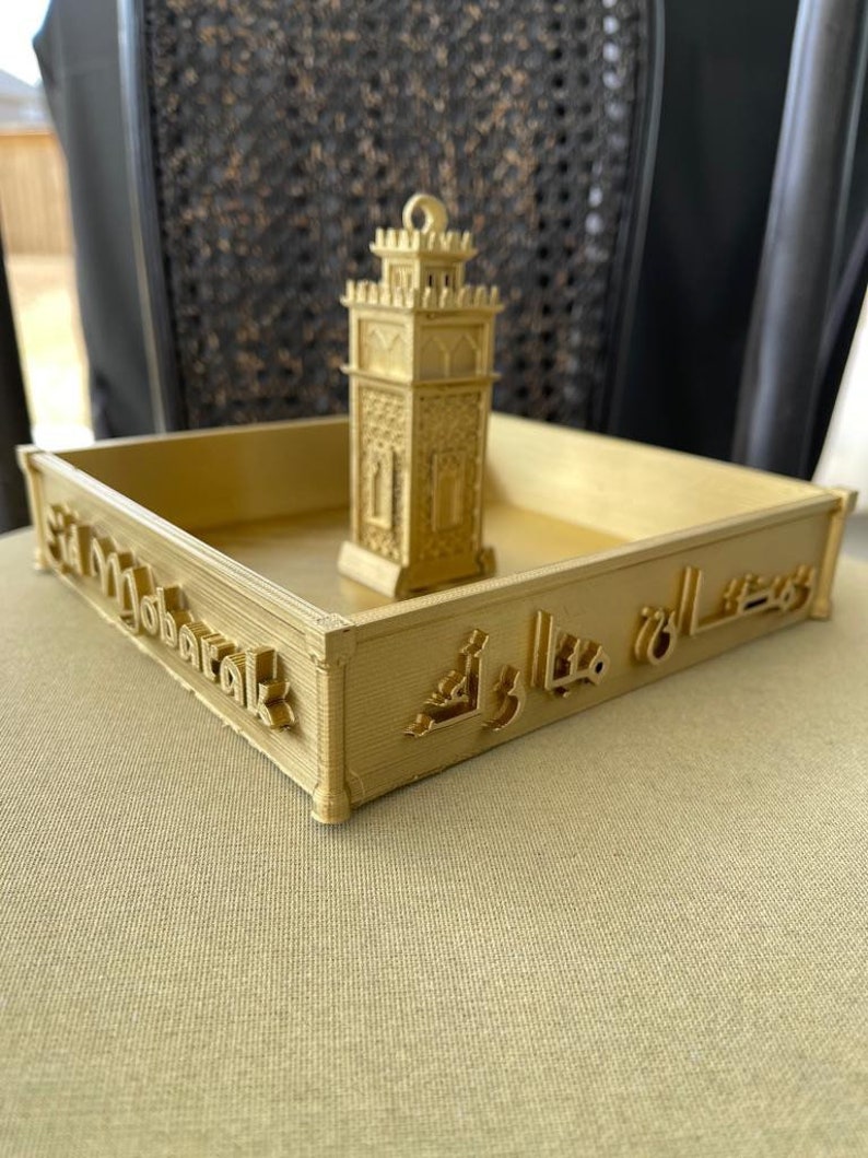 May include: A gold-colored tray with the words "Eid Mubarak" and Arabic script on the sides. A small gold-colored model of a minaret sits in the center of the tray.