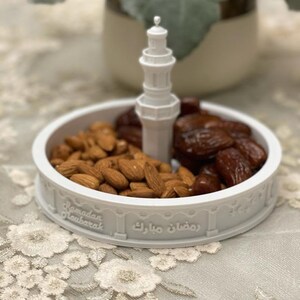 May include: White ceramic dish with a mosque-shaped design, featuring the text "Ramadan Mubarak" and "رمضان مبارك". The dish is filled with almonds and dates.