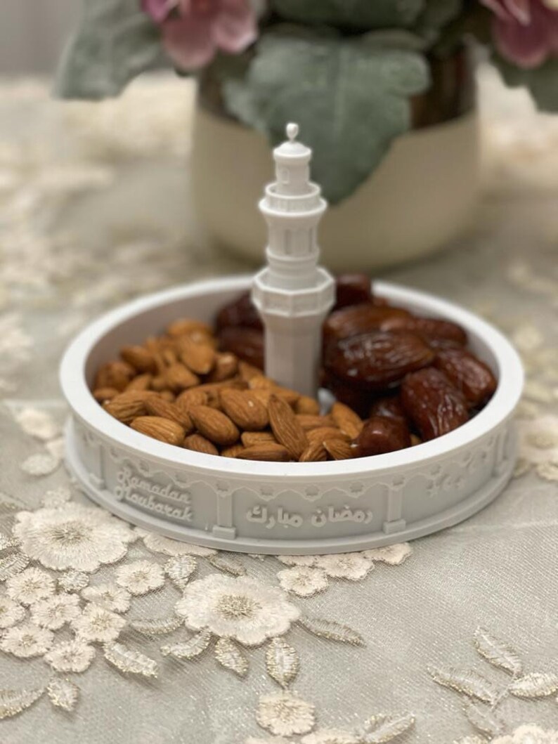 May include: White ceramic dish with a mosque-shaped design, featuring the text "Ramadan Mubarak" and "رمضان مبارك". The dish is filled with almonds and dates.