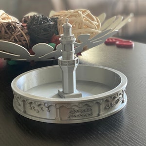 May include: A 3D printed silver model of a mosque with a minaret on a round base. The base has the words "Ramadan Mubarak" inscribed on it.