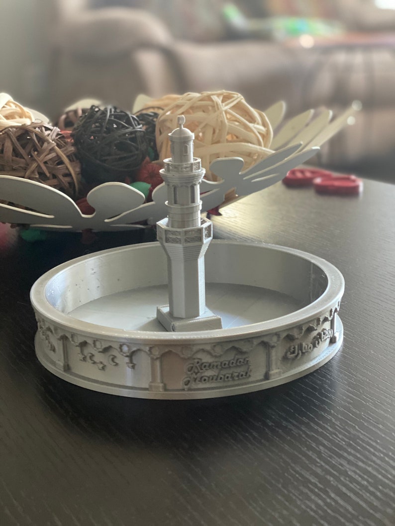 May include: A 3D printed silver model of a mosque with a minaret on a round base. The base has the words "Ramadan Mubarak" inscribed on it.