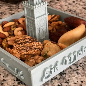 May include: A light blue tray with the words "Eid Mubarak" on the side. The tray is filled with nuts, dates, and other treats. A small, light blue 3D printed mosque sits in the center of the tray.