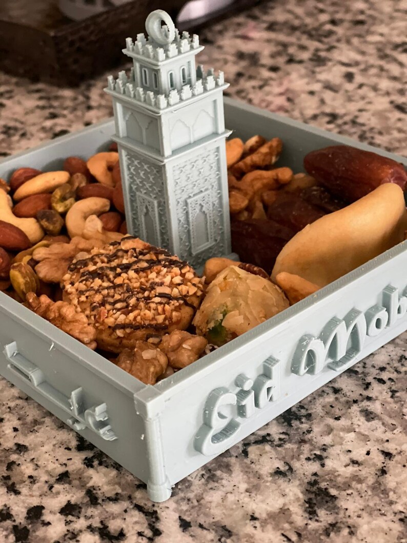 May include: A light blue tray with the words "Eid Mubarak" on the side. The tray is filled with nuts, dates, and other treats. A small, light blue 3D printed mosque sits in the center of the tray.