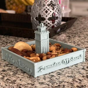 May include: A light blue ceramic dish shaped like a mosque with a small minaret on top. The dish is filled with nuts, dates, and other dried fruits. The dish has the text "Ramadan Mubarak" and Arabic text on the side.