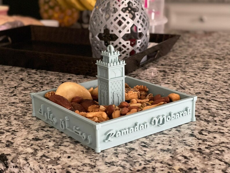 May include: A light blue ceramic dish shaped like a mosque with a small minaret on top. The dish is filled with nuts, dates, and other dried fruits. The dish has the text "Ramadan Mubarak" and Arabic text on the side.