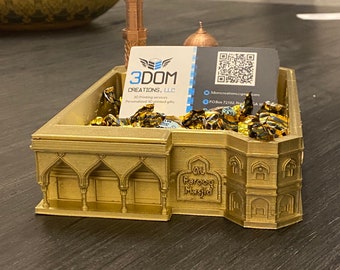 May include: A gold 3D printed model of a mosque with a minaret, a dome, and arches. The model is sitting on a table with a business card that says "3DOM Creations, LLC" and a QR code. The model is surrounded by gold-colored candy.