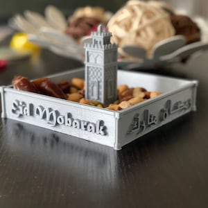 May include: A gray 3D printed tray with the words "Eid Mubarak" and Arabic script. The tray is filled with nuts and dates. A small gray 3D printed mosque is in the center of the tray.