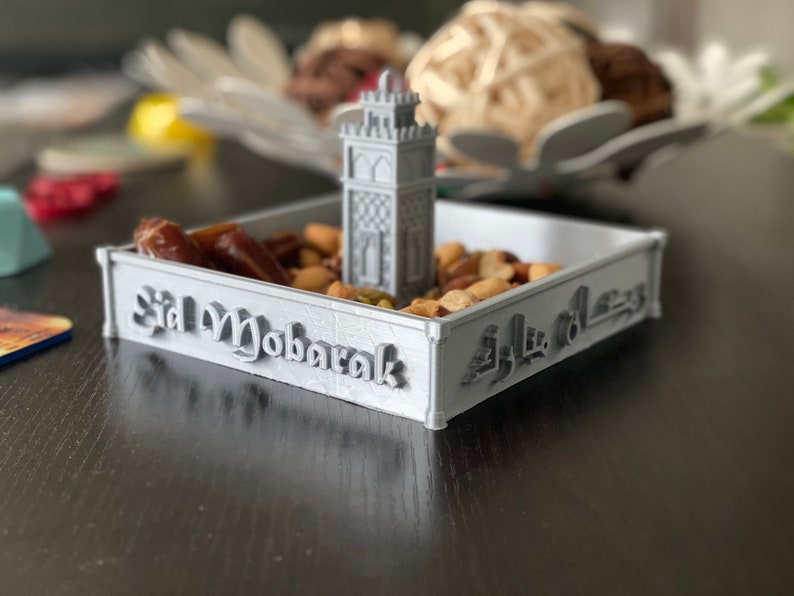 May include: A gray 3D printed tray with the words "Eid Mubarak" and Arabic script. The tray is filled with nuts and dates. A small gray 3D printed mosque is in the center of the tray.