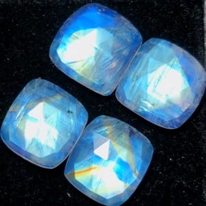 May include: Four square-cut, iridescent moonstone gemstones. Each stone displays a captivating play of blue and white light, with a faceted surface that catches and reflects light. The gemstones are set against a dark background.