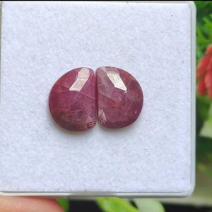 Peut inclure: Deux pierres précieuses rubis ovales et facettées. Les rubis sont d'un rouge profond et riche avec des veines internes. Elles sont présentées dans un récipient carré blanc, avec un arrière-plan vert et rouge flou.