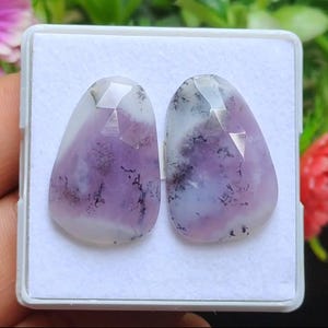 May include: Two polished gemstones with a mix of white, lavender, and purple hues. The stones have a faceted top and a teardrop shape, with dark, tree-like inclusions. They are set in a white box.