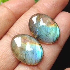 May include: Two oval-shaped labradorite gemstones. The stones display a mix of colors, including blue, green, and gold, with a shimmering, iridescent effect. The polished stones are smooth and have a glossy finish.