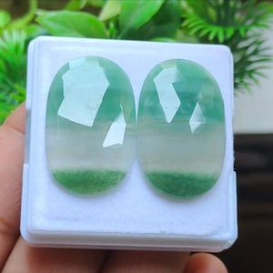 May include: Two oval gemstones with a faceted cut, presented in a white box. The stones display a colour gradient, shifting from clear to light green, with a vivid green base. These gemstones are likely for jewellery making.