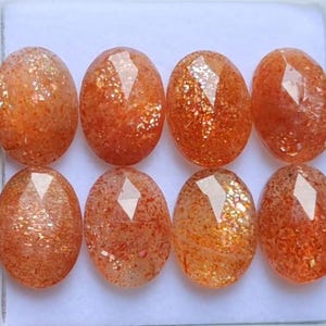Peut inclure: Huit pierres précieuses ovales en sunstone. Les pierres sont d'une couleur orange chaud avec des inclusions scintillantes. Certaines pierres ont des facettes. Les pierres sont présentées dans un plateau blanc.