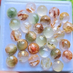 May include: A selection of faceted, spherical gemstones in shades of clear, green, brown, and yellow. The gemstones are presented in a white tray, highlighting their polished surfaces and internal features. These gems are likely for jewellery making.
