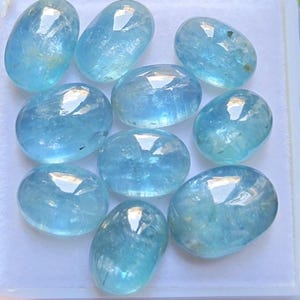 May include: A collection of nine oval-shaped, polished light blue gemstones. Each stone features a smooth, cabochon-style surface with translucent qualities and internal inclusions, arranged in a cluster on a white display tray.