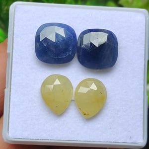 Op de afbeelding: Vier edelstenen gepresenteerd in een witte tray. Twee zijn kussenvormige, diepblauwe saffieren. De andere twee zijn peervormige, lichtgele saffieren. De edelstenen hebben gefacetteerde oppervlakken die het licht reflecteren.