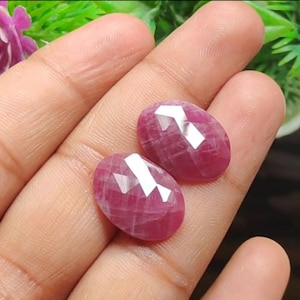 May include: Two faceted, oval-shaped rubies. The gemstones are a deep red color with bright, reflective facets. The rubies are held in a hand, with a blurred background of green foliage.