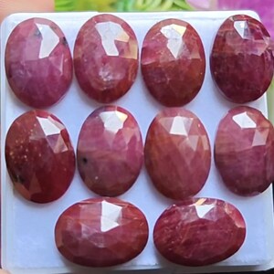 Puede incluir: Una colección de nueve rubíes de corte ovalado, cada uno mostrando un tono rojo intenso con superficies facetadas. Las piedras preciosas están dispuestas en una cuadrícula, mostrando su apariencia pulida y su potencial para el diseño de joyas. Los rubíes varían ligeramente en color y claridad.